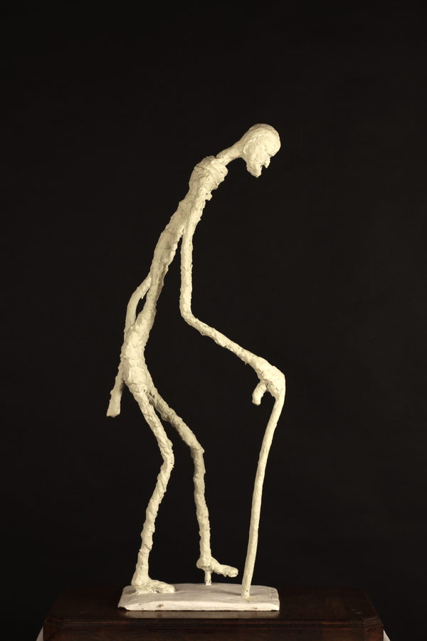 Old Man Walking (2020) by Valeriu Cazacevschi. A pale, elongated old man bends forward over a walking stick, on a dark wooden base.