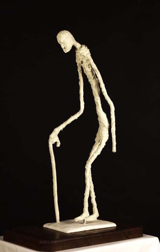 Pale elongated figure leaning forward on a walking stick