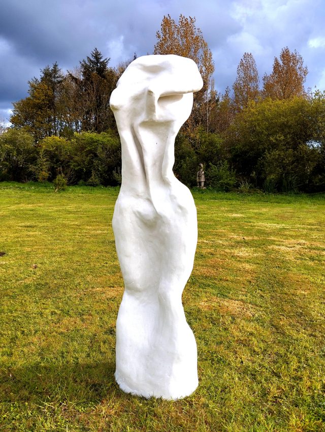 Tall white figurative sculpture photographed outdoors in a garden